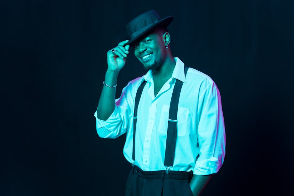 Confident black man in stylish attire with suspenders, tipping his hat and smiling on black background.