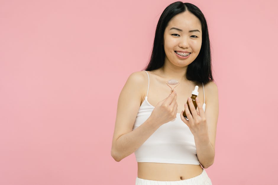 Woman smiling with facial roller and serum bottle against a pink backdrop, promoting skincare.