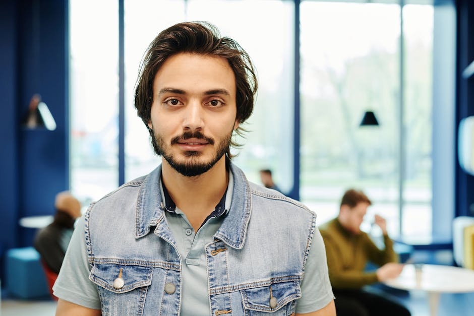 Relaxed portrait of a bearded man in a denim vest inside a modern cafe.
