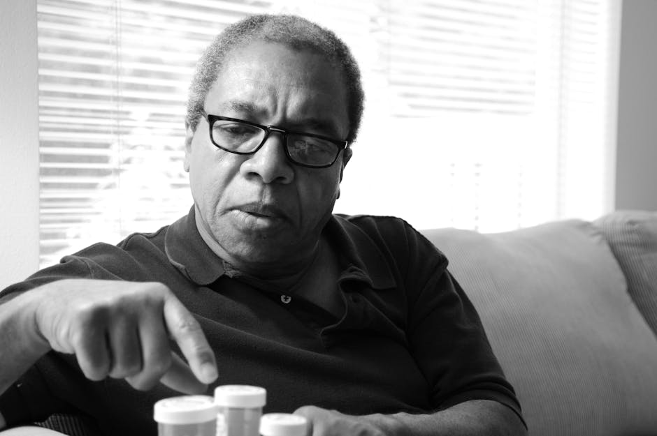 Black and white photo of an adult man sitting indoors with medication bottles.