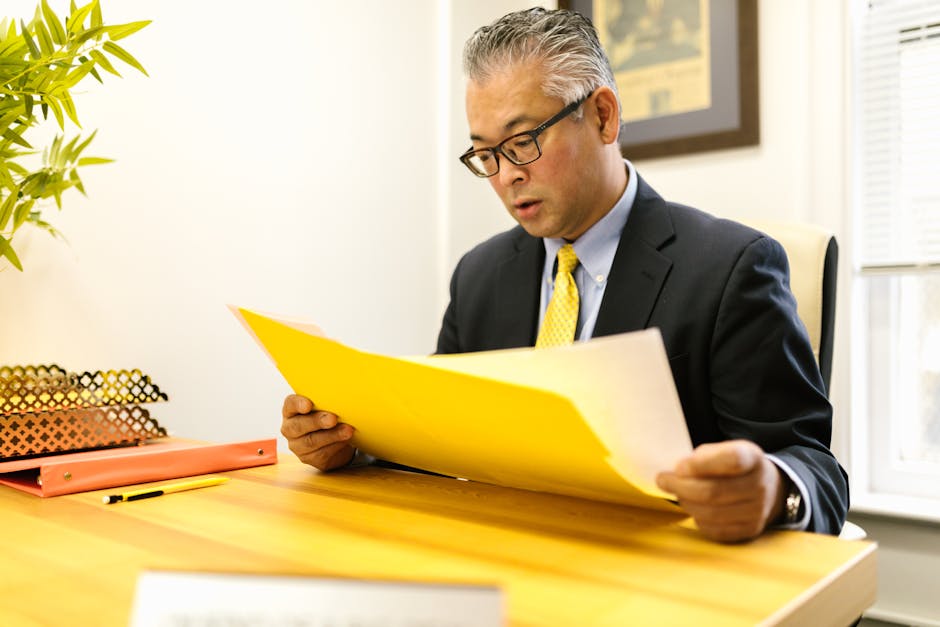 Asian businessman in corporate attire reading documents at office desk with a yellow folder.