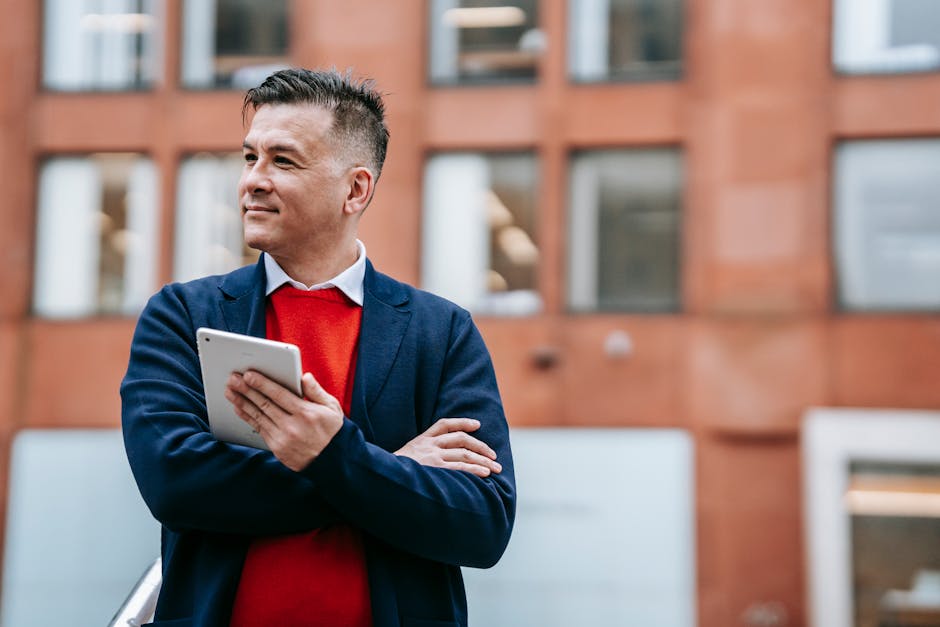 Smiling businessman in a blazer holding a tablet, standing outside an office building.
