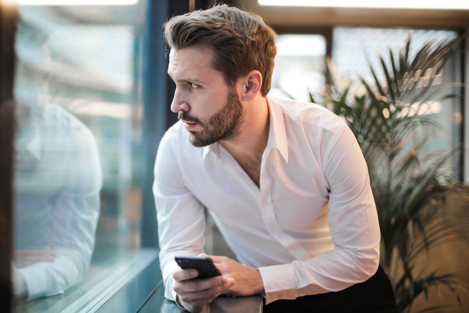 A thoughtful man with a smartphone looking out an office window.