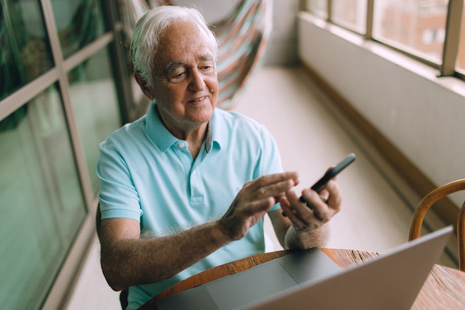 Elderly man in blue polo shirt using smartphone and laptop indoors.