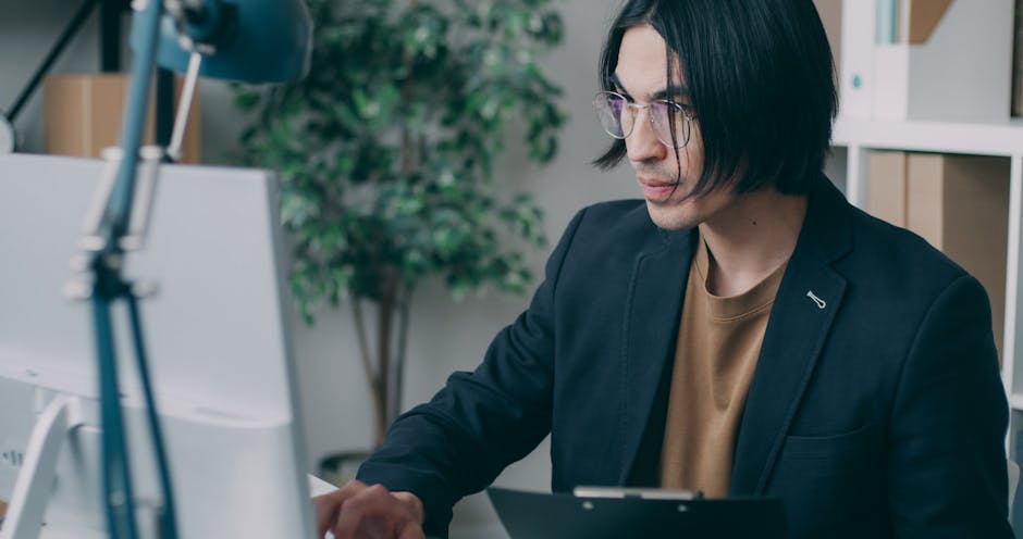 Man in glasses concentrating on work at a computer desk in a contemporary office setting.