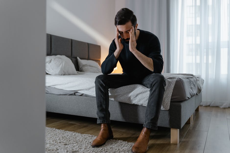 Adult man sitting on bed, appearing stressed or frustrated, holding his temples in a well-lit bedroom.